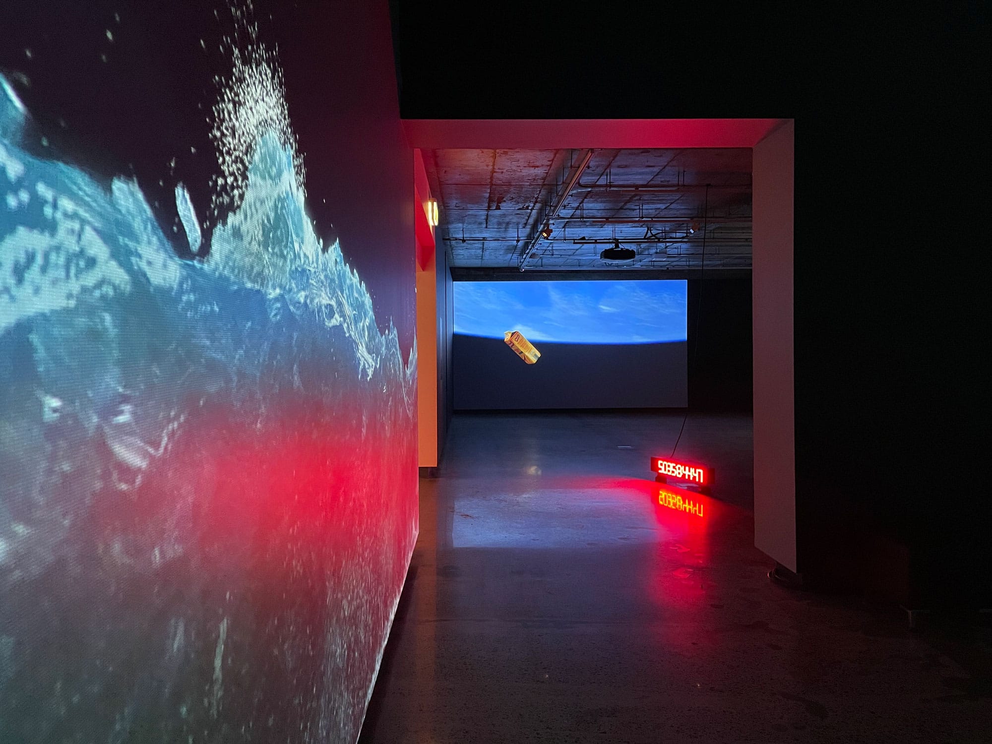Projection art in the NorthSite Gallery by Yandell Walton. A wave is on the left wall and on the back wall is a juice bottle floating in the water. A countdown time is in the midde illuminating the walls in red.