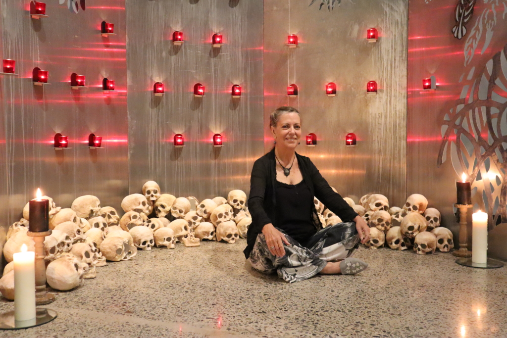 Artist Elizabeth Hunter sits in front of her shrine cross-legged with her hands on her knees. She is wearing a black top and black cardigan with patterned black and white pants. The shrine behind her has a large pile of hand-made sculls and candles in red candle jars light up the polished steel walls that form the shine. Larger tall candles are placed on the floor at either side of the shrine. 