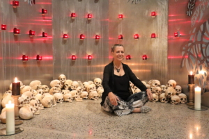 Artist Elizabeth Hunter sits in front of her shrine cross-legged with her hands on her knees. She is wearing a black top and black cardigan with patterned black and white pants. The shrine behind her has a large pile of hand-made sculls and candles in red candle jars light up the polished steel walls that form the shine. Larger tall candles are placed on the floor at either side of the shrine.