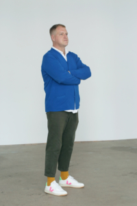Hamish Sawyer standing at a 45 degree angle with arms crossed. He is wearing a blue cardigan with a white collared shirt underneath. Charcoal chinos, orange socks and white shoes.