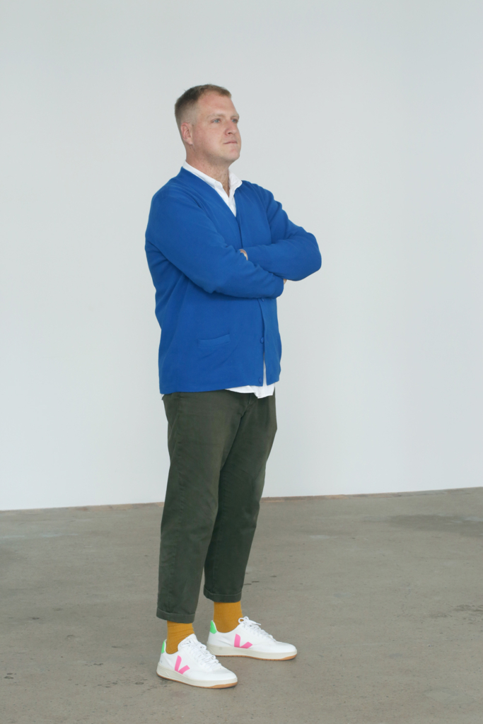 Hamish Sawyer standing at a 45 degree angle with arms crossed. He is wearing a blue cardigan with a white collared shirt underneath. Charcoal chinos, orange socks and white shoes. 