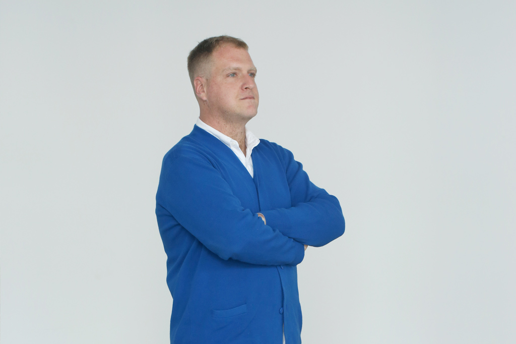 Hamish Sawyer standing at a 45 degree angle with arms crossed. He is wearing a blue cardigan with a white collared shirt underneath.