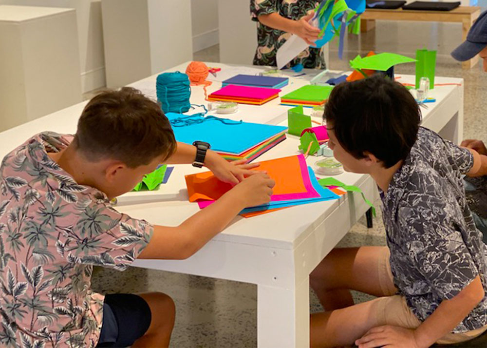 Two young boys sitting at a white table full of colourful paper and creative materials.