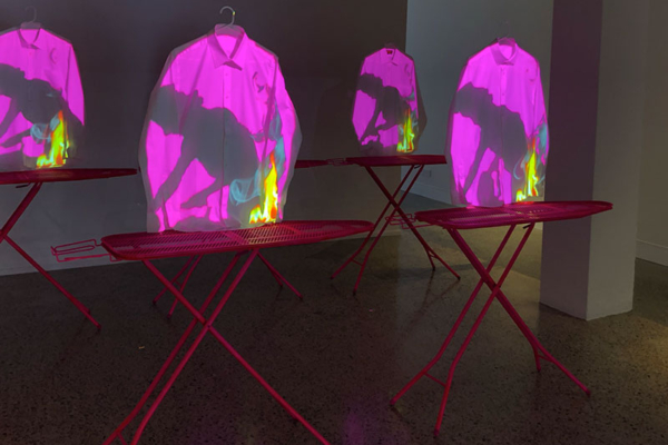 5 ironing boards, 2 in the front row and 3 in the back row, painted in bright pink. White collared shirts are suspended above each ironing board. Projected onto the white dress shirts is the silhouette of a 1950s quintessential housewife ironing.