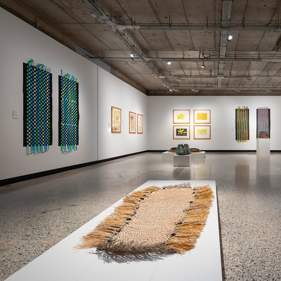 A gallery with white walls, polished concrete floor and concrete ceiling. A woven mat is in the foreground on a low white plinth. Artworks can be seen hanging on the walls around the gallery space.