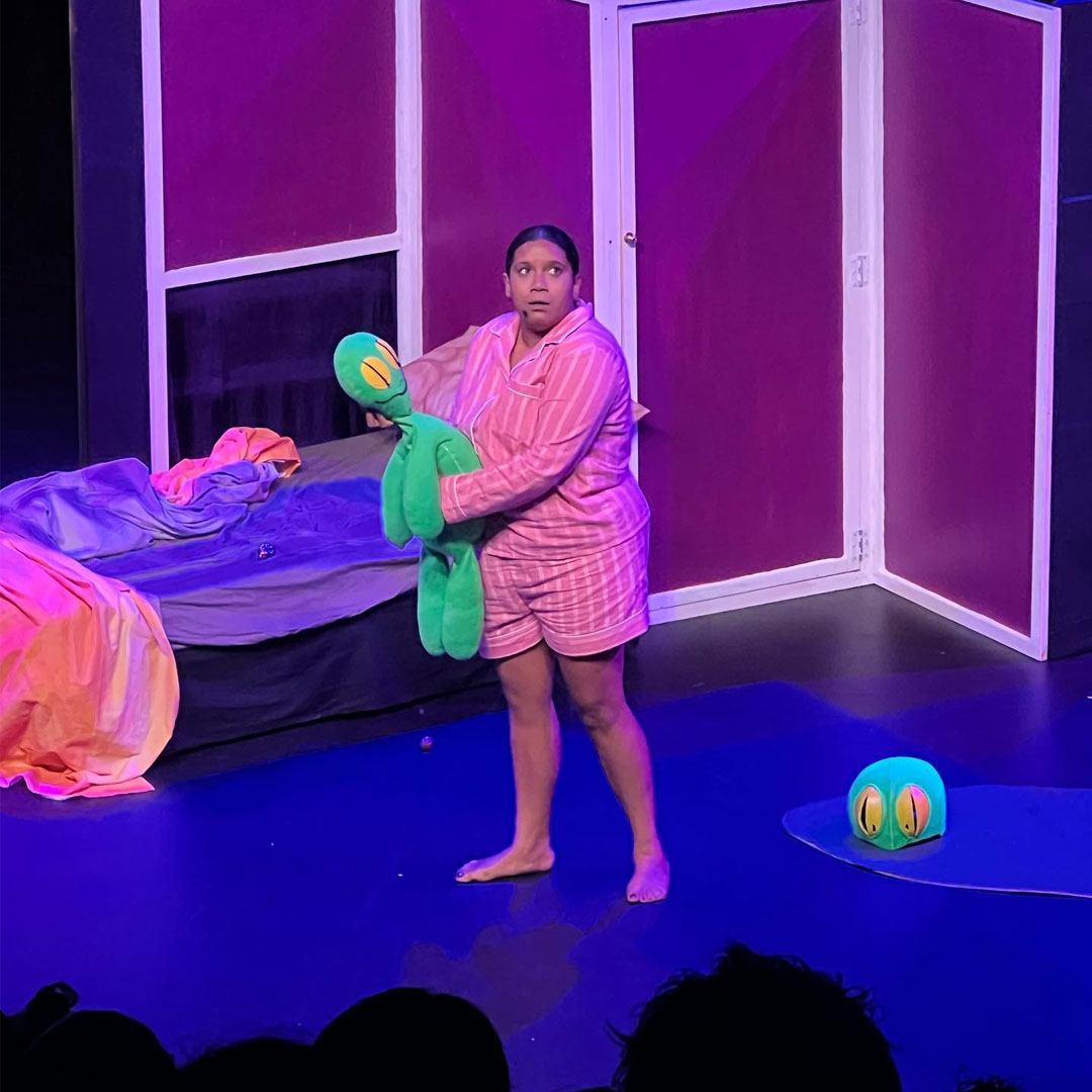 Performance: Young girl (played by Jamaylya Ballangarry-Kearins) stands holding her green toy gecko. She looks concerned. She is wearing a pink striped pyjama set.