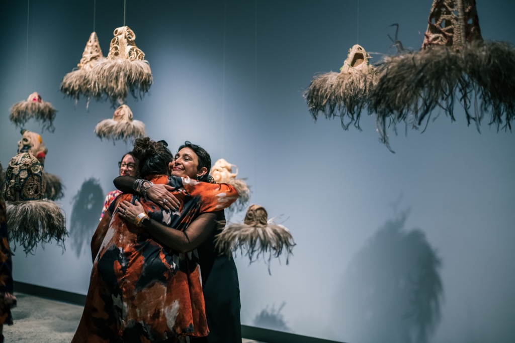 Two women hug in the gallery surrounded by woven sculptures hung from the double height ceiling. 