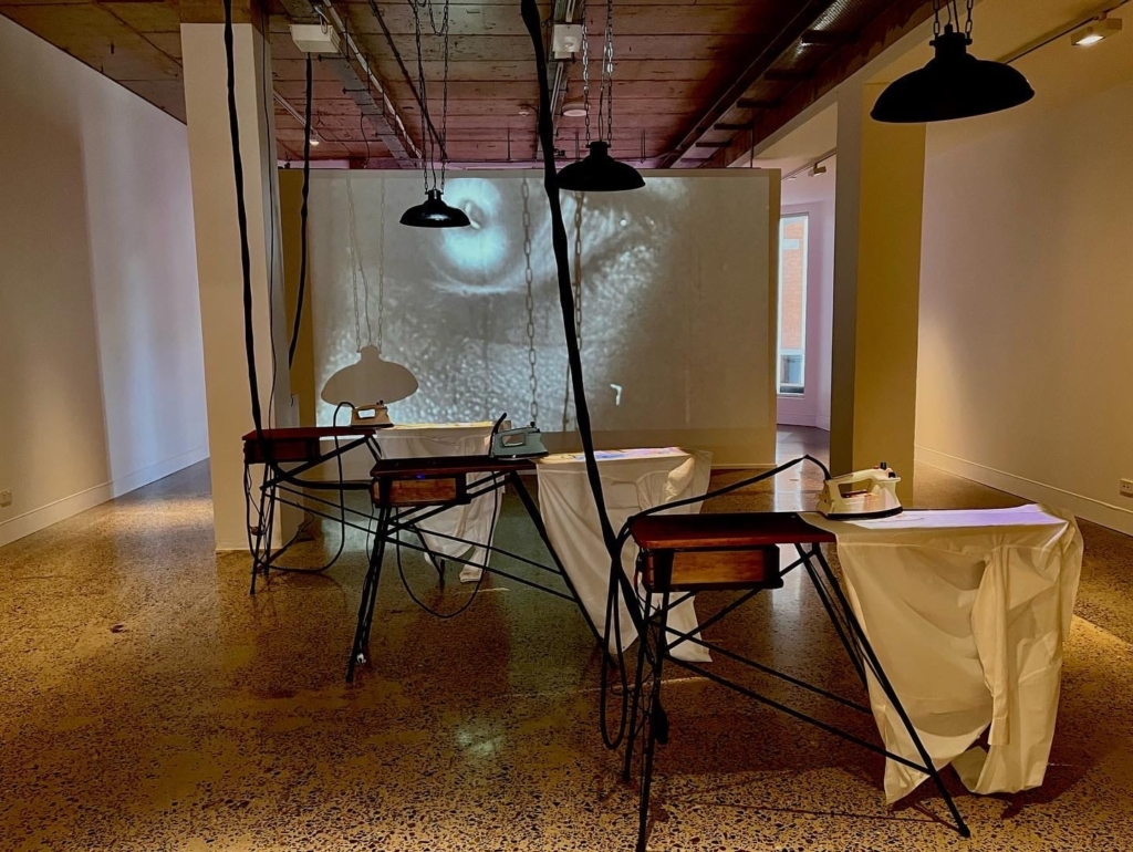 Installation of 3 ironing boards with retro irons. In the background is a video projection. Hanging from the ceiling are industrial lights and cords referencing the factories.