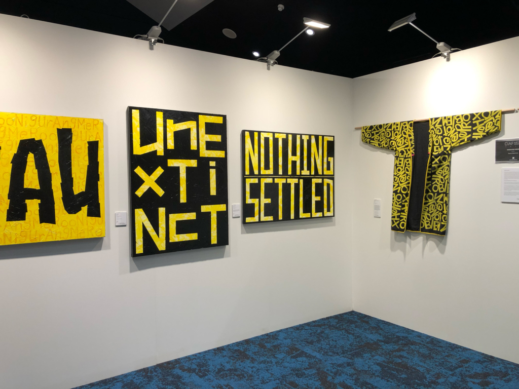 Four artworks by Darren Blackman hang on a gallery wall. All art black and yellow with text.