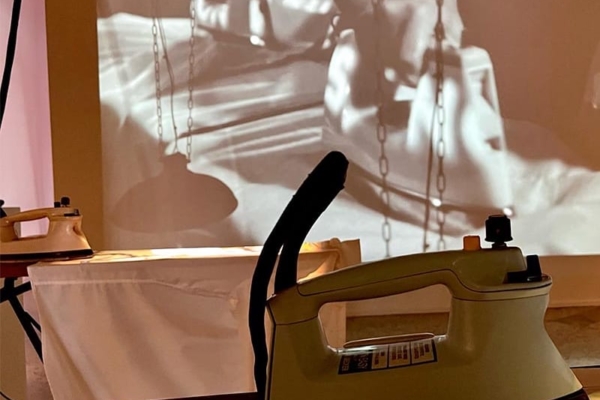 An old retro iron sitting on an ironing board. In the background is the projection with video people ironing in a factory.