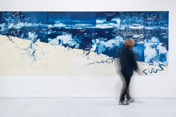 A large artwork on a white wall. The artwork is a detailed cyanotype on paper. A woman is blurred in the foreground walking towards the artwork and looking at it.