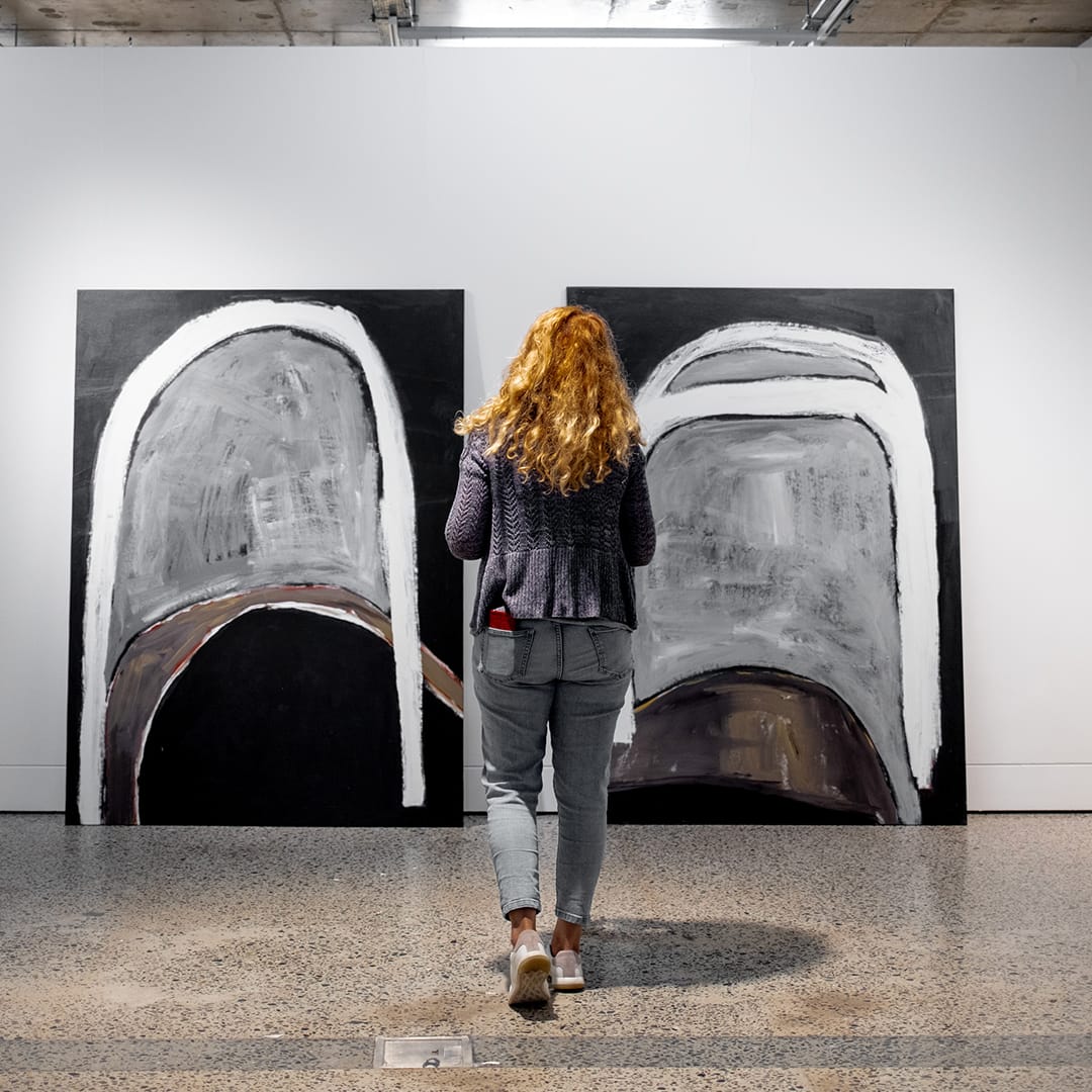 A woman walking away from the camera. There are two large artworks leaning against a white wall in the background that she is walking towards.