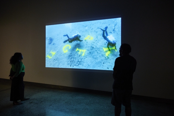 Two people standing in a room looking at a projection of scuba divers. In the still image of the film, there are several letters 'n, g, f, p, r' that the divers are moving around the ocean floor.