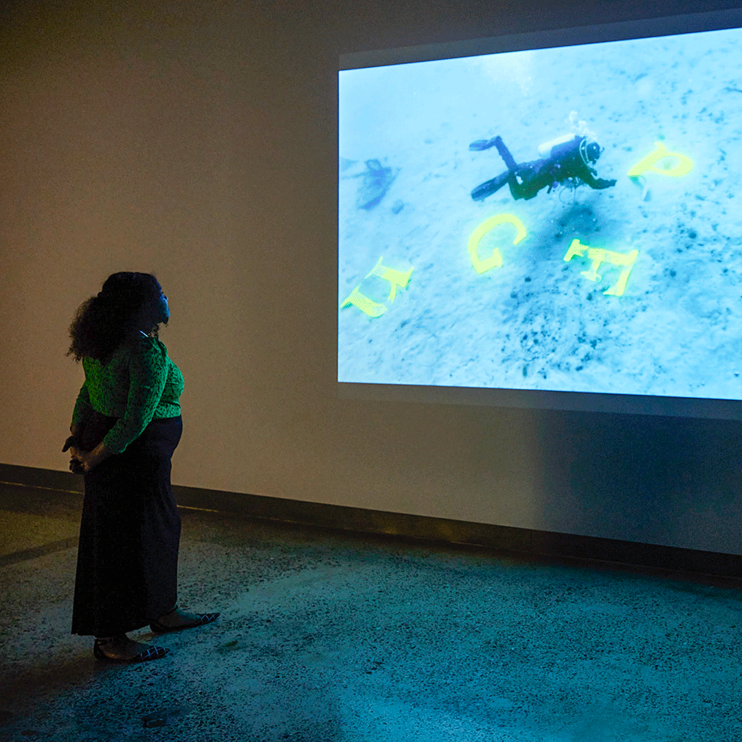 A woman standing in a room looking at a projection of scuba divers. In the still image of the film, there are several letters 'n, g, f, p, r' that the divers are moving around the ocean floor.