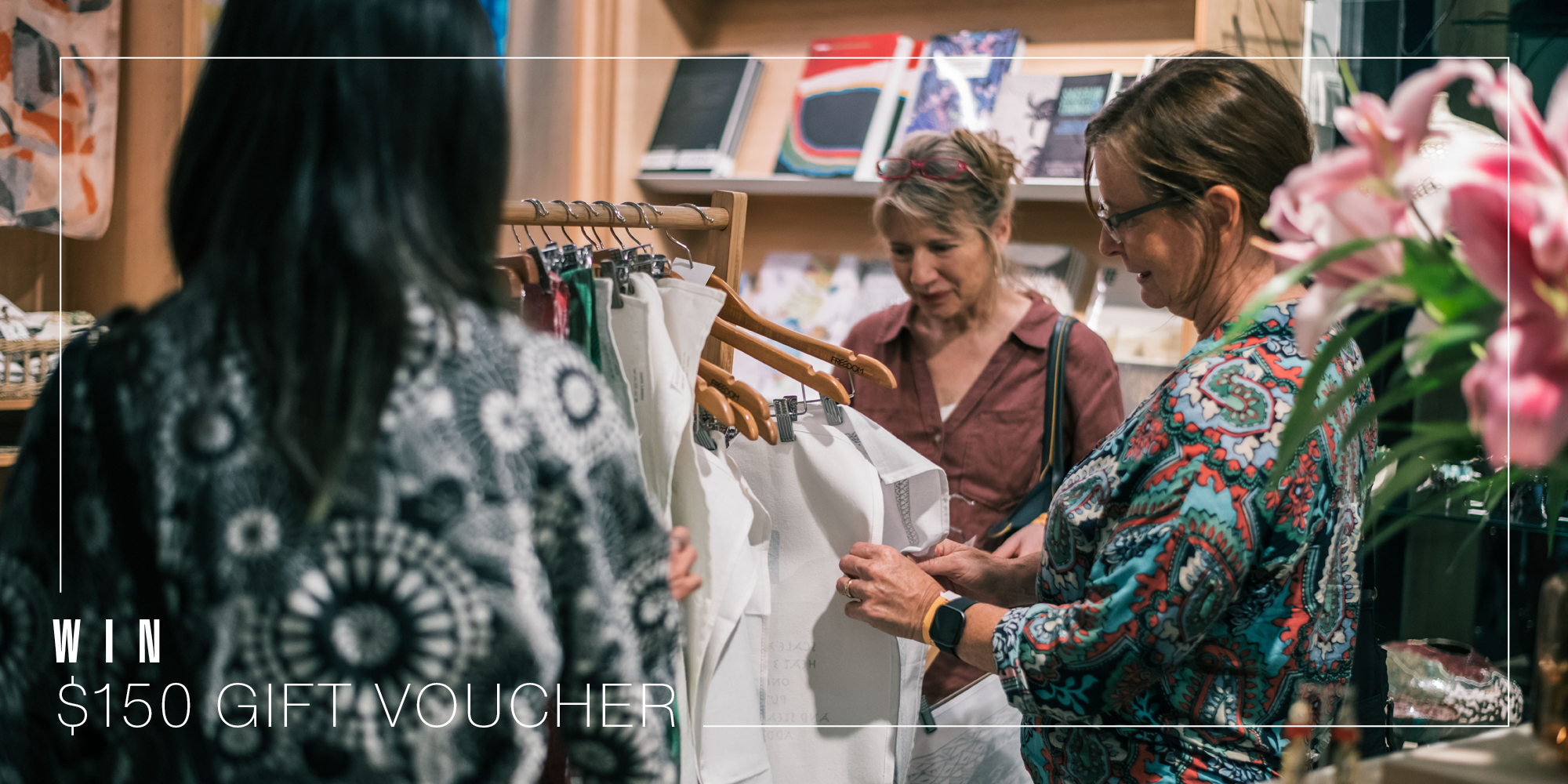 Three women looking at items in the NorthSite Store. Pink lilies are in the right corner. A thin white line frames the image with the text 'Win a $150 Gift Vourcher'.