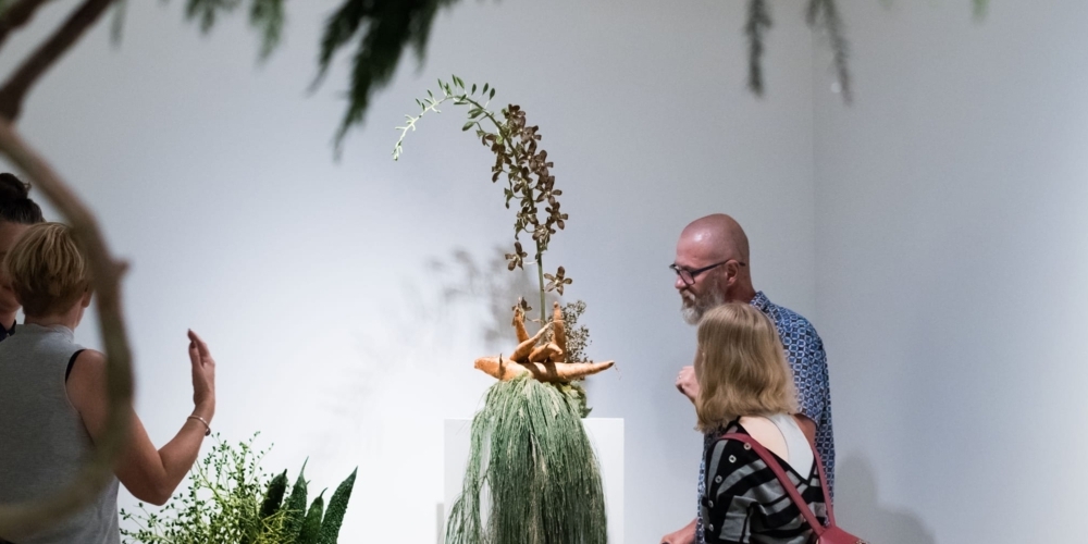 Four people are walking around the gallery space full of plants. Two arrangements are presented on two tall plinths and a larger sculptural piece is in the foreground.