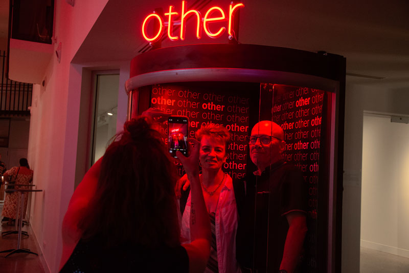 Two people get their photo taken inside a booth with a neon light that reads 'other'