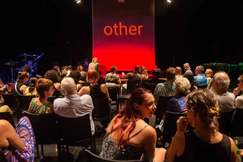 An image of a room of people sitting and chatting in the crowd. In the distance is a stage lit in red lights and the text 'other'.