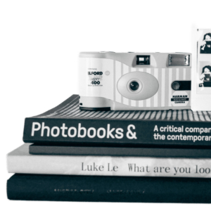 A black and white photograph of 4 books stacked and a camera and Polaroid on-top of the books. 